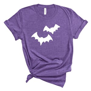 May include: Purple t-shirt with two white bat silhouettes. The short-sleeved shirt is knotted at the bottom. The shirt is made of a soft material and is suitable for Halloween or everyday wear.