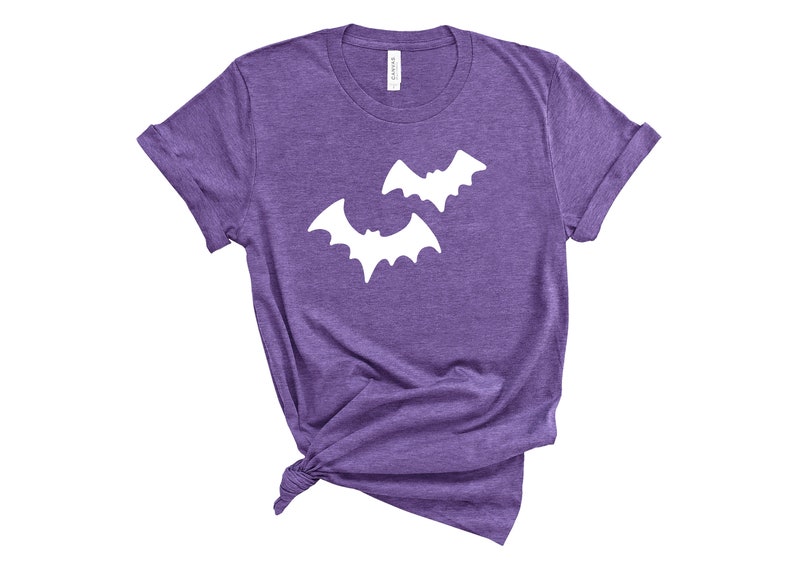 May include: Purple t-shirt with two white bat silhouettes. The short-sleeved shirt is knotted at the bottom. The shirt is made of a soft material and is suitable for Halloween or everyday wear.