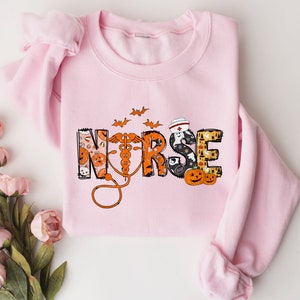 May include: A light pink sweatshirt with the word "NURSE" in a Halloween-themed design. The letters are decorated with pumpkins, bats, and a nurse's cap. An orange stethoscope is featured.
