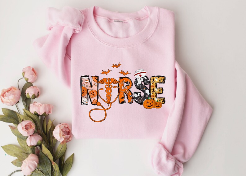May include: A light pink sweatshirt with the word "NURSE" in a Halloween-themed design. The letters are decorated with pumpkins, bats, and a nurse's cap. An orange stethoscope is featured.