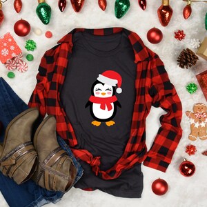 May include: A black t-shirt with a cute cartoon penguin wearing a red Santa hat and scarf. The shirt is laid out on a white surface with red and green Christmas decorations.
