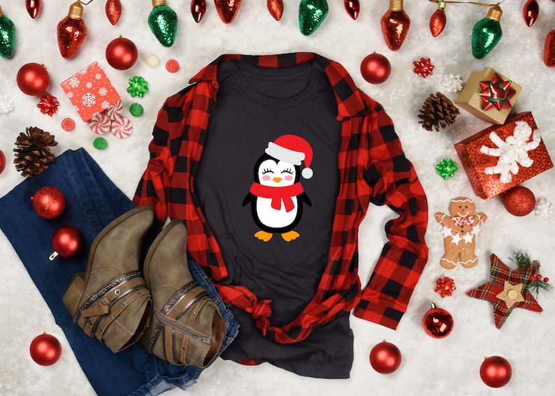 May include: A black t-shirt with a cute cartoon penguin wearing a red Santa hat and scarf. The shirt is laid out on a white surface with red and green Christmas decorations.