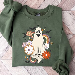 May include: An olive green sweatshirt with a smiling ghost design, flowers, bats, a rainbow, and a crescent moon. The graphic has a retro, vintage style, suitable for Halloween or autumn. The long-sleeved top is made of a soft material.
