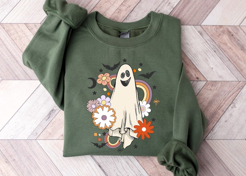 May include: An olive green sweatshirt with a smiling ghost design, flowers, bats, a rainbow, and a crescent moon. The graphic has a retro, vintage style, suitable for Halloween or autumn. The long-sleeved top is made of a soft material.