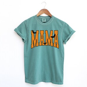May include: A teal-colored t-shirt with the word "MAMA" in orange block letters. Black bat silhouettes are placed above and below the word "MAMA". The shirt is hanging on a wooden hanger.