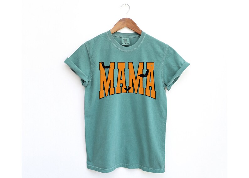 May include: A teal-colored t-shirt with the word "MAMA" in orange block letters. Black bat silhouettes are placed above and below the word "MAMA". The shirt is hanging on a wooden hanger.