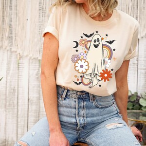 May include: Cream-coloured t-shirt with a retro ghost design, including a rainbow, flowers, bats, and stars. The ghost has a smiling face. This casual tee is ideal for Halloween or the spooky season.