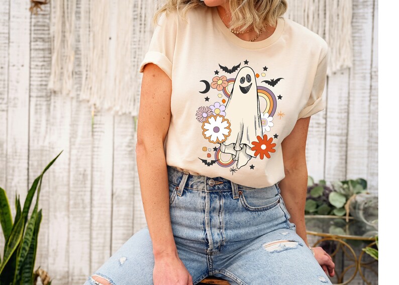 May include: Cream-coloured t-shirt with a retro ghost design, including a rainbow, flowers, bats, and stars. The ghost has a smiling face. This casual tee is ideal for Halloween or the spooky season.