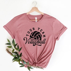 Puede incluir: Una camiseta rosa con un gr&aacute;fico de voleibol negro y el texto "Your Team Volleyball" impreso en la parte delantera.