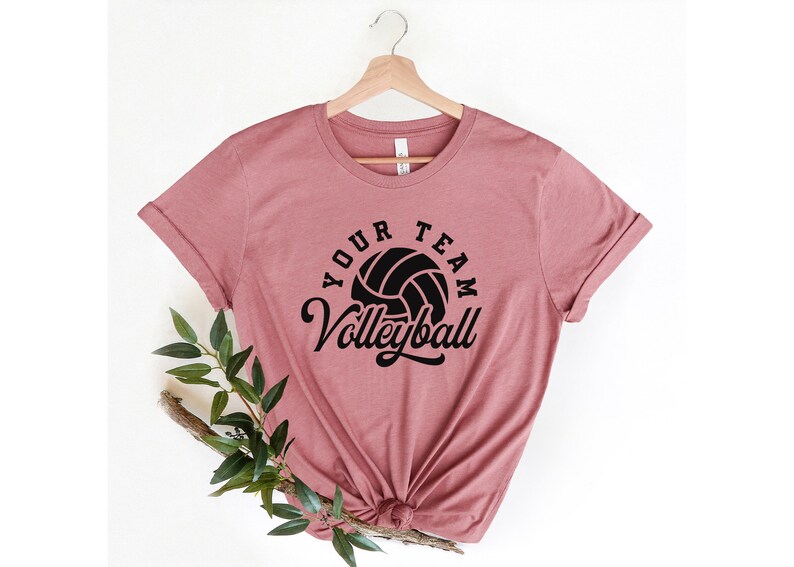 Puede incluir: Una camiseta rosa con un gr&aacute;fico de voleibol negro y el texto "Your Team Volleyball" impreso en la parte delantera.