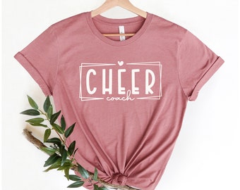 May include: A dusty rose t-shirt with white text that reads "CHEER coach" with a small heart above the word "CHEER".