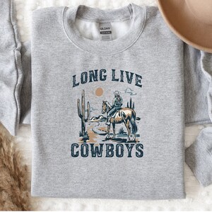 May include: A gray sweatshirt with a graphic print of a cowboy riding a horse in a desert landscape. The text "Long Live Cowboys" is printed on the sweatshirt.