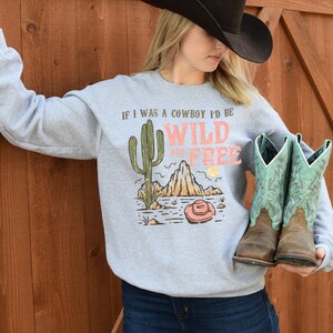 May include: Gray sweatshirt with a graphic print of a cactus, mountains, and a cowboy hat. The text on the shirt reads "If I was a cowboy I'd be wild and free".