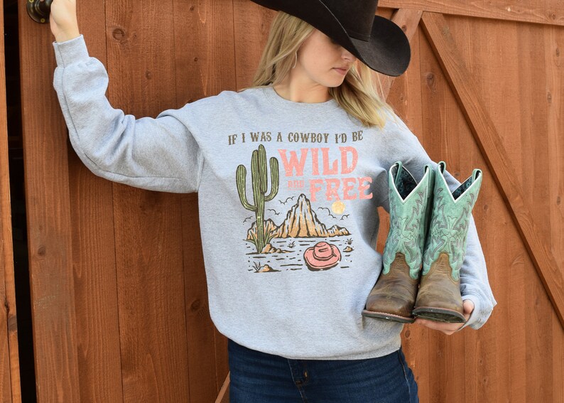 May include: Gray sweatshirt with a graphic print of a cactus, mountains, and a cowboy hat. The text on the shirt reads "If I was a cowboy I'd be wild and free".