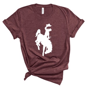 May include: A maroon t-shirt with a white silhouette of a cowboy riding a bucking horse.