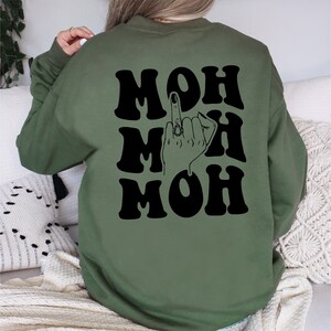Pode incluir: Camisola verde com texto preto que diz "MOH MOH MOH" e uma ilustra&ccedil;&atilde;o preta de uma m&atilde;o a fazer o dedo do meio com um anel no dedo do meio.