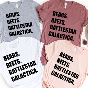 May include: Four t-shirts in gray, maroon, white, and peach, each with the text "BEARS. BEETS. BATTLESTAR GALACTICA." in black. Sunglasses and passports are visible in the background.