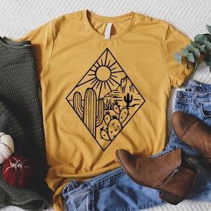K&ouml;nnte beinhalten: Ein senfgelbes T-Shirt mit einer schwarzen Grafik einer W&uuml;stenlandschaft mit Sonne, Kaktus und Bergen. Das T-Shirt liegt auf einer wei&szlig;en Oberfl&auml;che mit einer blauen Jeans und braunen Stiefeletten.