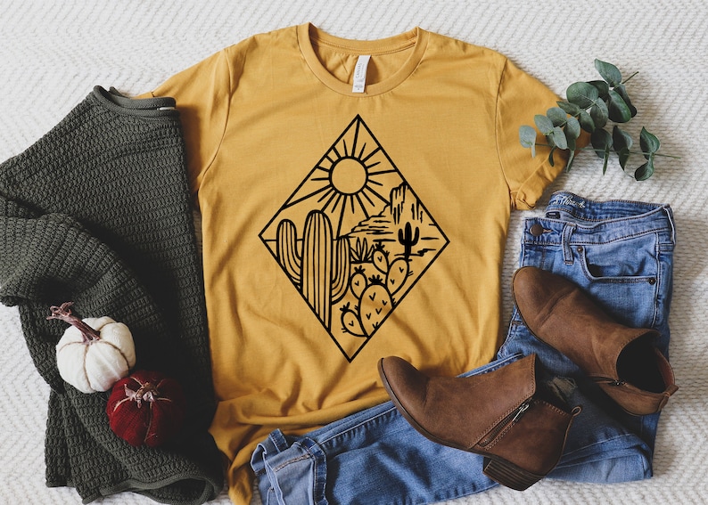K&ouml;nnte beinhalten: Ein senfgelbes T-Shirt mit einer schwarzen Grafik einer W&uuml;stenlandschaft mit Sonne, Kaktus und Bergen. Das T-Shirt liegt auf einer wei&szlig;en Oberfl&auml;che mit einer blauen Jeans und braunen Stiefeletten.