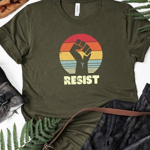 May include: A green t-shirt with a vintage-style graphic of a raised fist in front of a sunset. The text "RESIST" is printed below the graphic.