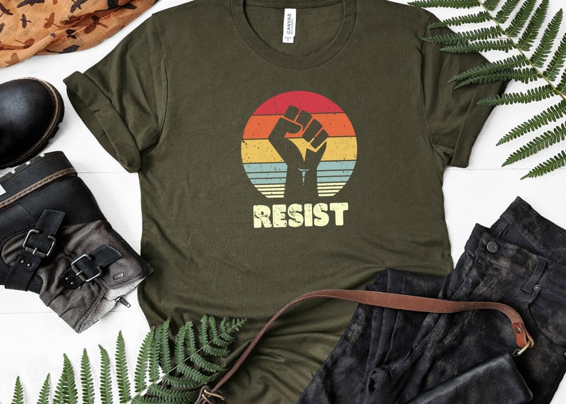 May include: A green t-shirt with a vintage-style graphic of a raised fist in front of a sunset. The text "RESIST" is printed below the graphic.
