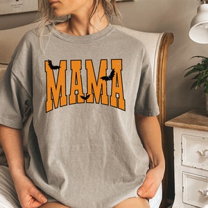 May include: A gray t-shirt with the word "MAMA" in orange block letters, outlined in black. Black bat silhouettes are above and below the word, creating a Halloween theme. The shirt is a casual, oversized style.