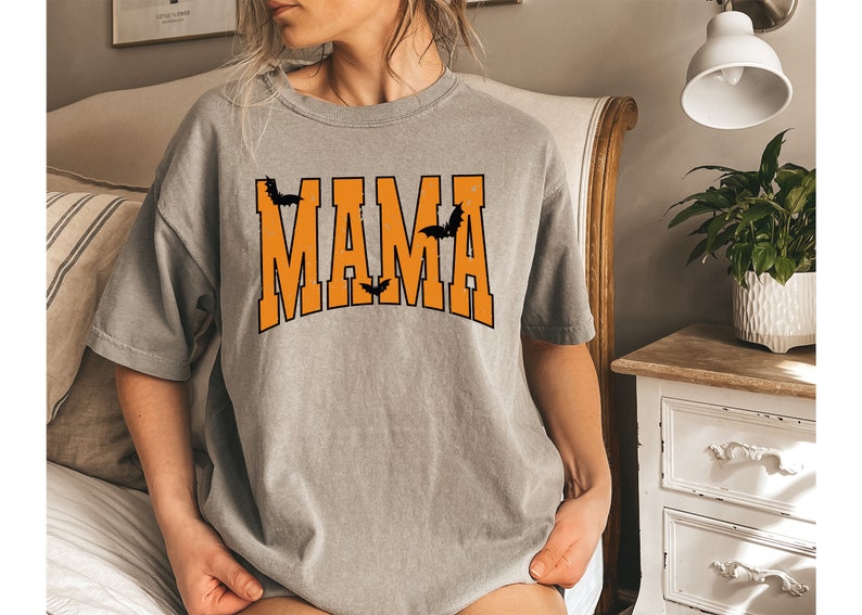 May include: A gray t-shirt with the word "MAMA" in orange block letters, outlined in black. Black bat silhouettes are above and below the word, creating a Halloween theme. The shirt is a casual, oversized style.