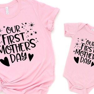 May include: Pink t-shirt and onesie set with black text that reads "Our First Mother's Day" with stars and hearts.