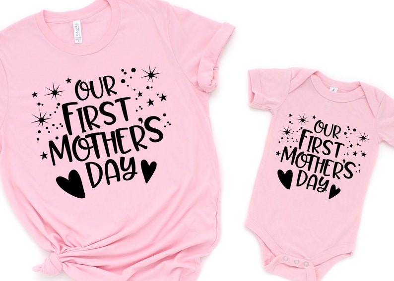 May include: Pink t-shirt and onesie set with black text that reads "Our First Mother's Day" with stars and hearts.