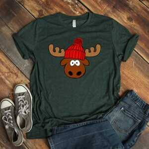 May include: A dark green t-shirt with a cartoon moose wearing a red knit hat with a white pom-pom.