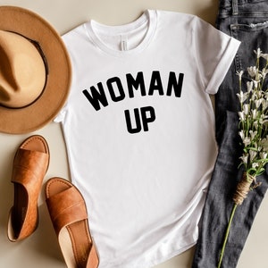 May include: White t-shirt with black text that reads "WOMAN UP" on a white background. The shirt is laid flat on a light brown surface with a brown hat, brown sandals, and a bouquet of white flowers and dark blue jeans.