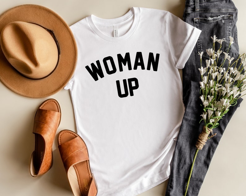 May include: White t-shirt with black text that reads "WOMAN UP" on a white background. The shirt is laid flat on a light brown surface with a brown hat, brown sandals, and a bouquet of white flowers and dark blue jeans.