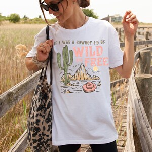 May include: White t-shirt with a graphic print of a cactus, a mountain, and a cowboy hat. The text on the shirt reads "If I was a cowboy I'd be Wild and Free".