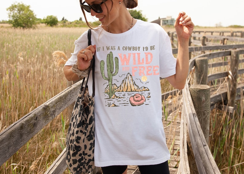 May include: White t-shirt with a graphic print of a cactus, a mountain, and a cowboy hat. The text on the shirt reads "If I was a cowboy I'd be Wild and Free".