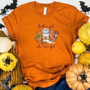 May include: Orange t-shirt with the text "Just a girl who loves fall" and a fall-themed graphic. The graphic includes a coffee cup, boots, pumpkins, and an apple. Decorative gourds, spiders, and bats surround the shirt.