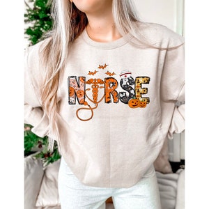 May include: Beige sweatshirt with the word "NURSE" in a Halloween-themed design. The letters are decorated with pumpkins, bats, and a medical symbol. The sweatshirt is a crew neck style.