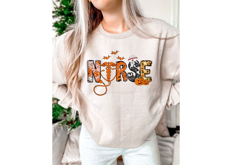 May include: Beige sweatshirt with the word "NURSE" in a Halloween-themed design. The letters are decorated with pumpkins, bats, and a medical symbol. The sweatshirt is a crew neck style.