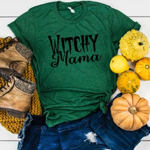 May include: A green t-shirt with the text "Witchy Mama" in black script. The shirt is paired with denim shorts, brown boots, and a mustard-colored knit scarf. Decorative gourds in various colors and shapes are arranged around the shirt.