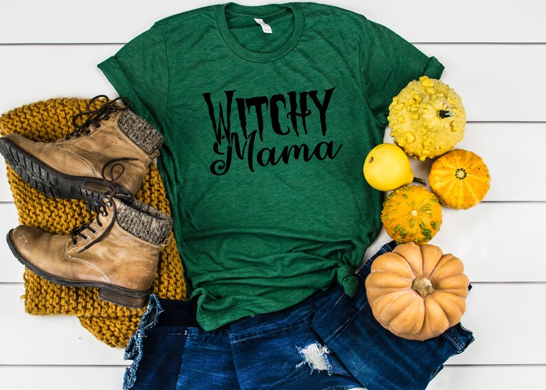 May include: A green t-shirt with the text "Witchy Mama" in black script. The shirt is paired with denim shorts, brown boots, and a mustard-colored knit scarf. Decorative gourds in various colors and shapes are arranged around the shirt.