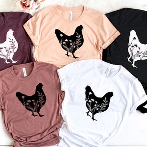 Pode incluir: Cinco camisetas com uma silhueta preta de uma galinha com flores dentro da silhueta. As camisetas s&atilde;o de v&aacute;rias cores: vinho, p&ecirc;ssego, preto, marrom e branco.