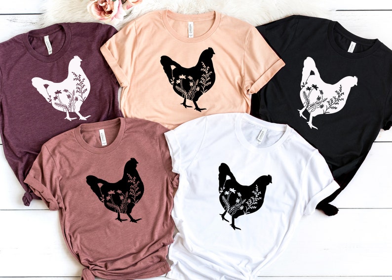 Pode incluir: Cinco camisetas com uma silhueta preta de uma galinha com flores dentro da silhueta. As camisetas s&atilde;o de v&aacute;rias cores: vinho, p&ecirc;ssego, preto, marrom e branco.