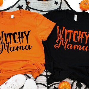 May include: Two t-shirts, one orange and one black, with the text "Witchy Mama" in a spooky font. The shirts are displayed with Halloween decorations, including pumpkins and a spiderweb.