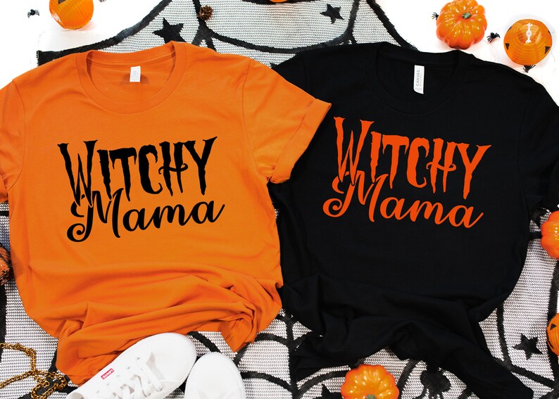 May include: Two t-shirts, one orange and one black, with the text "Witchy Mama" in a spooky font. The shirts are displayed with Halloween decorations, including pumpkins and a spiderweb.