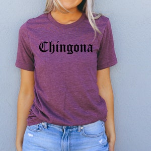 May include: A purple t-shirt with the word "Chingona" printed in black gothic lettering.