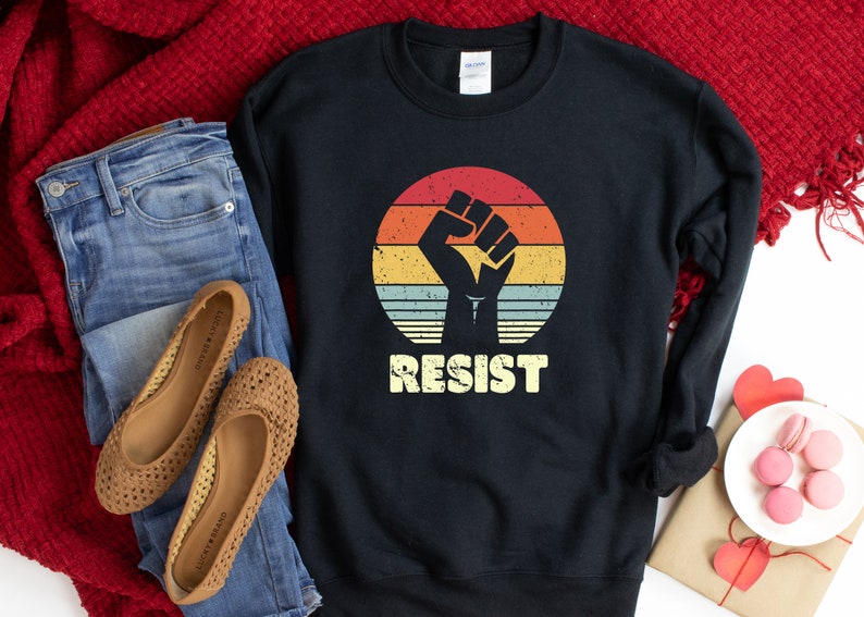 May include: Black sweatshirt with a vintage-style graphic of a raised fist in front of a sunset. The text "RESIST" is printed below the graphic.