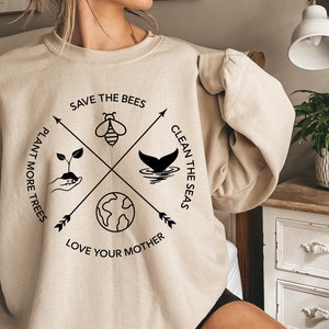 Może przedstawiać: Beżowa bluza z czarnym nadrukiem graficznym przedstawiającym pszczołę, ogon wieloryba, kiełek i glob. Tekst głosi "Save the Bees", "Plant More Trees", "Clean the Seas" i "Love Your Mother".