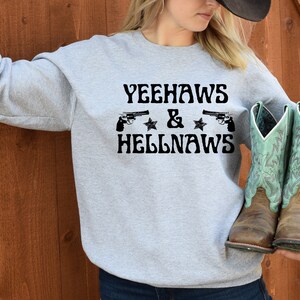 May include: Gray sweatshirt with the text "Yeehaws & Hellnaws" printed on the front. Two black pistols are shown above the text. The person is wearing blue jeans and holding a pair of brown and turquoise cowboy boots.