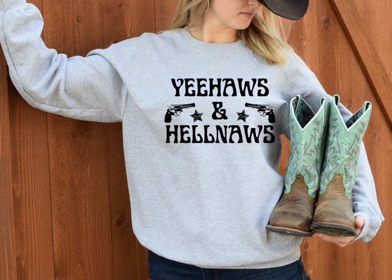 May include: Gray sweatshirt with the text "Yeehaws & Hellnaws" printed on the front. Two black pistols are shown above the text. The person is wearing blue jeans and holding a pair of brown and turquoise cowboy boots.