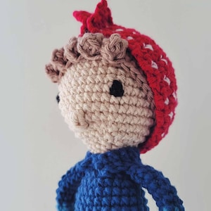 May include: A handmade crochet doll with a light beige face, brown hair, and a red hat with white accents. The doll wears a blue shirt with darker blue sleeves. The doll's eyes are black.