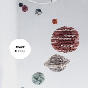 May include: A crocheted space mobile with planets in various colors, including red, gray, blue, and green. The mobile is hanging from a white circular base and has the text "SPACE MOBILE" on a white circle.